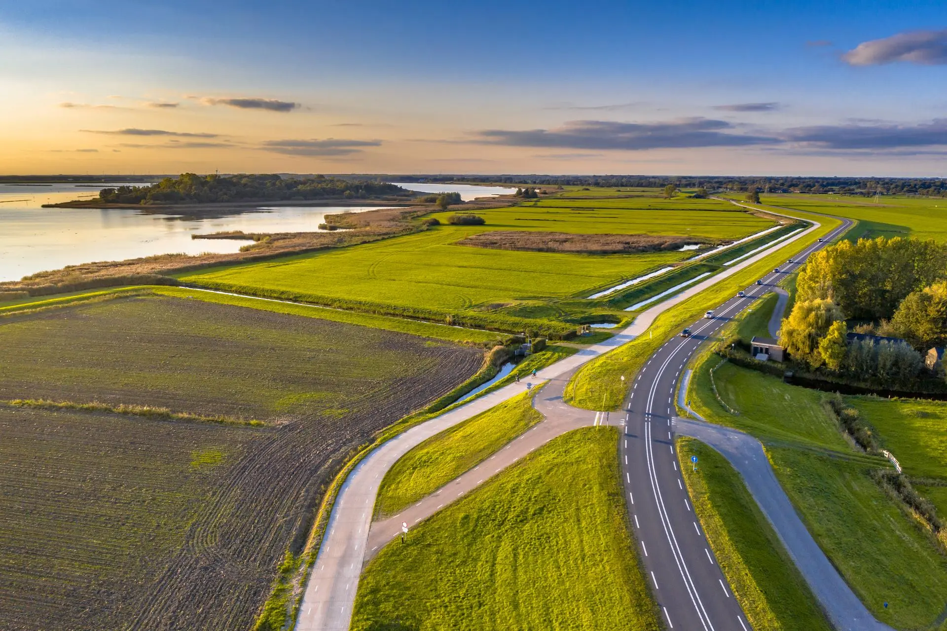 A road along a dyke through green polder fields beside the IJsselmeer, at sunrise