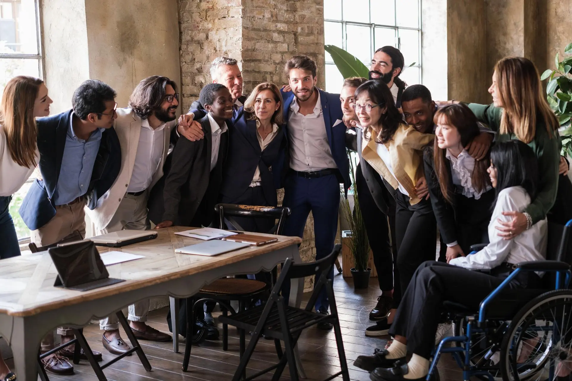 A group of colleagues of different ages and backgrounds stand close together around a table, one in a wheelchair