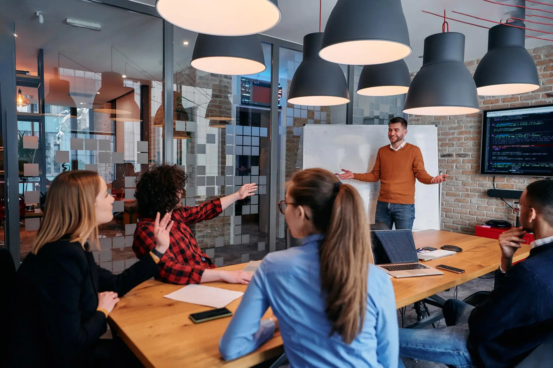 A trainer at a whiteboard leads a session; a small group around a table listens and responds